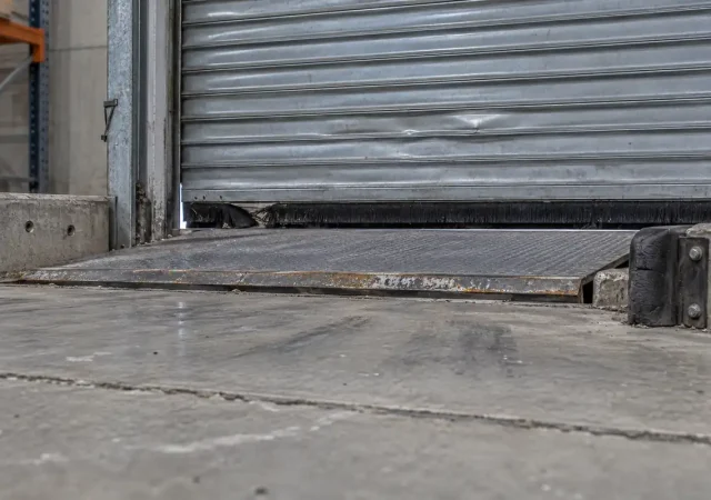 The inside foot of a closed warehouse roller shutter with a worn brush seal, a dock leveller plate and a daylight gap showing at the guide channel, warehouse proofing