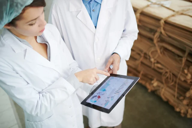 Two technologists in white coats reviewing pest monitoring data on a tablet beside stacked sacks in a food store